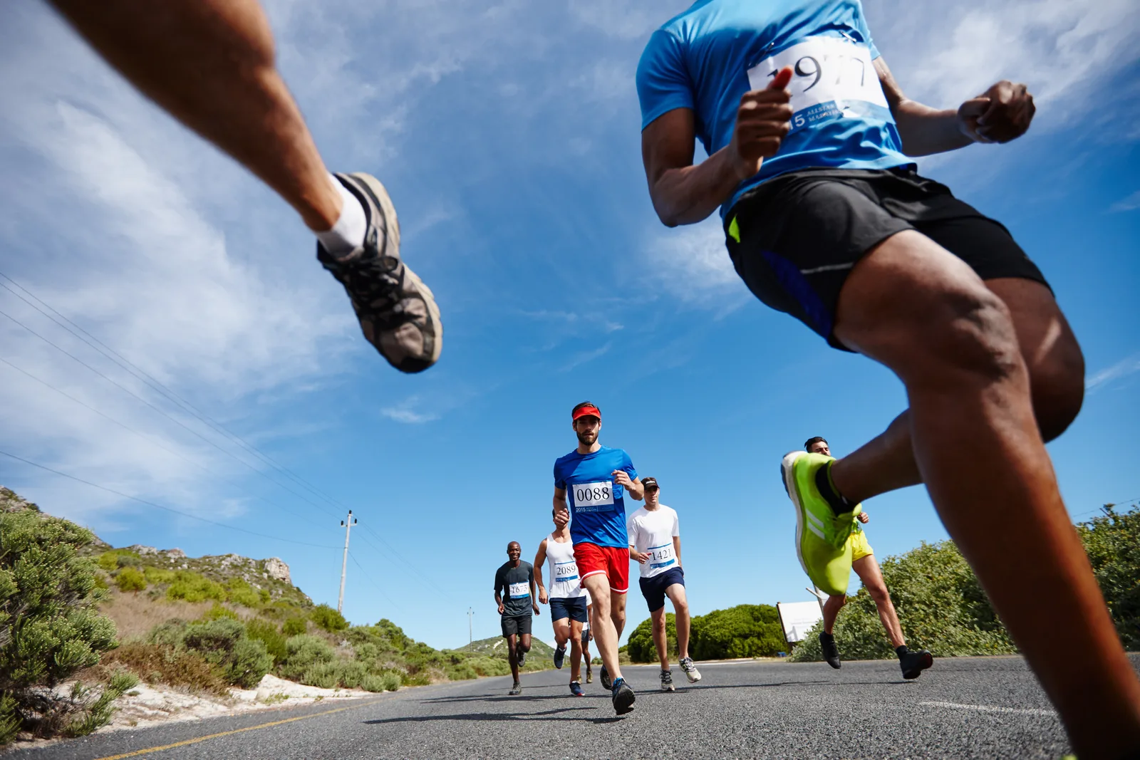 A tight pack of runners in shorts and singlets on an open road, seen low from ahead, arms swinging and every one of them mid-stride
